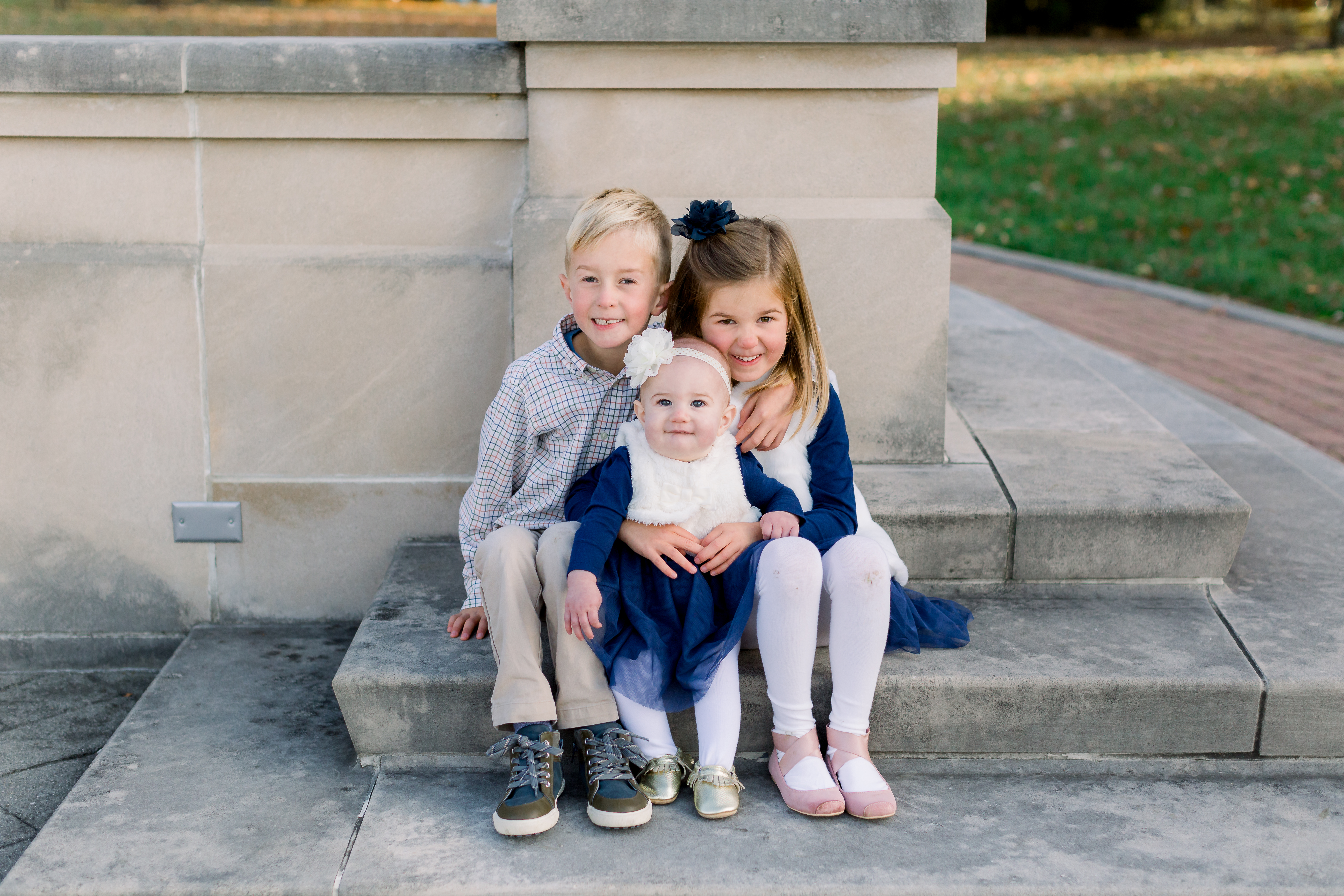 Three little kids cuddling together for a picture sitting on white concrete at Coxhall Gardens.