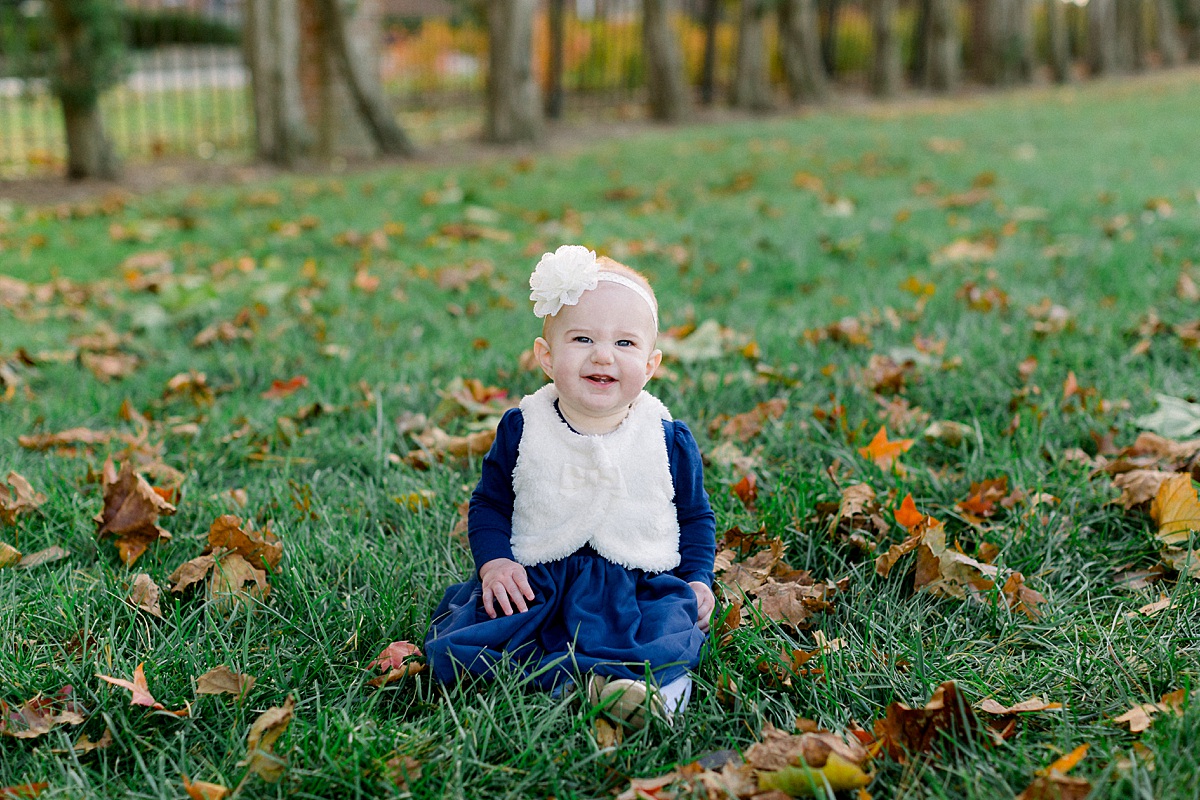Baby girl sitting in the grass with fall leaves all around her smiling for family photos. 