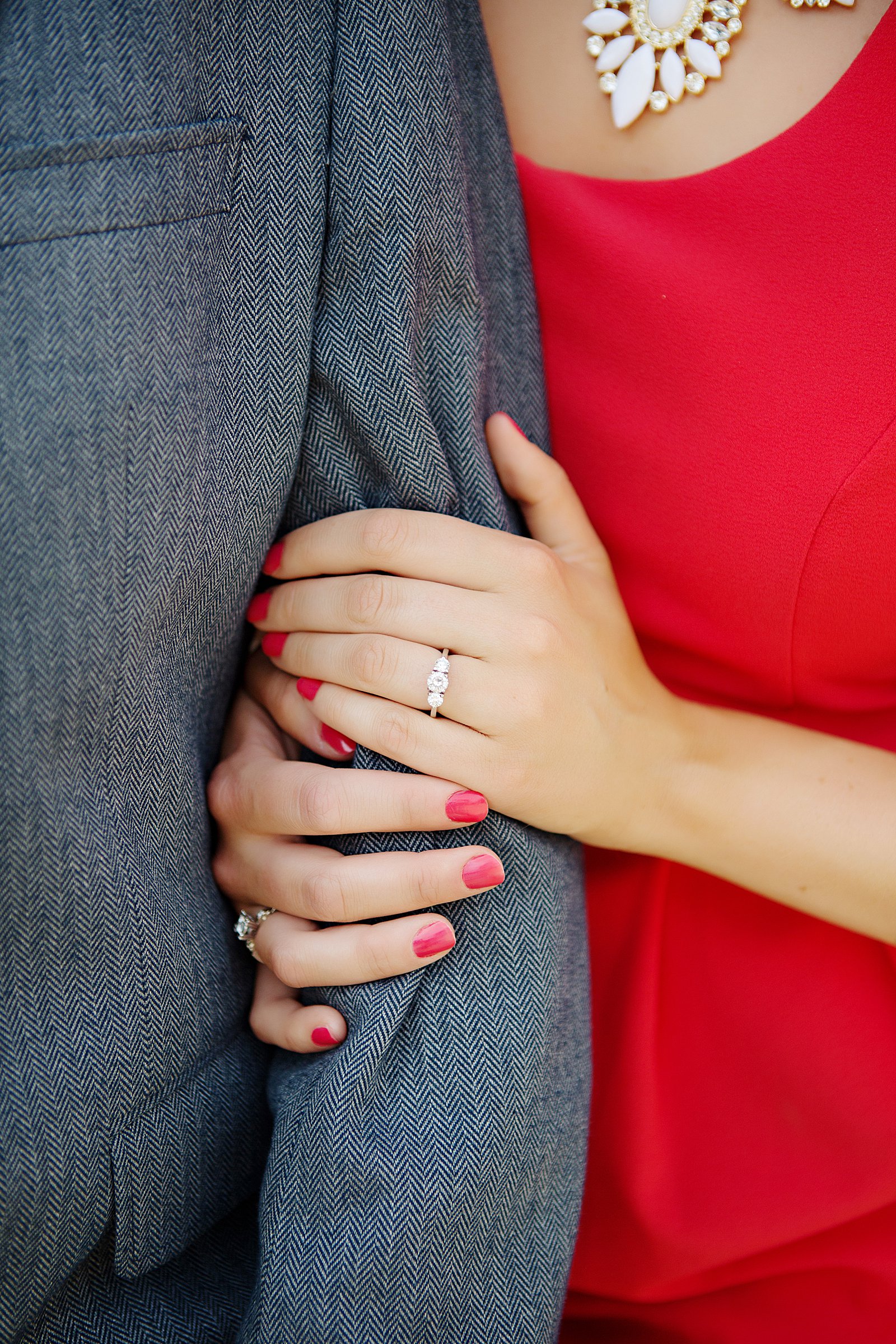Close up picture of a girls engagement ring with red nails and a red dress on. She is holding the arm of her fiancé at University of Illinois for photos. 