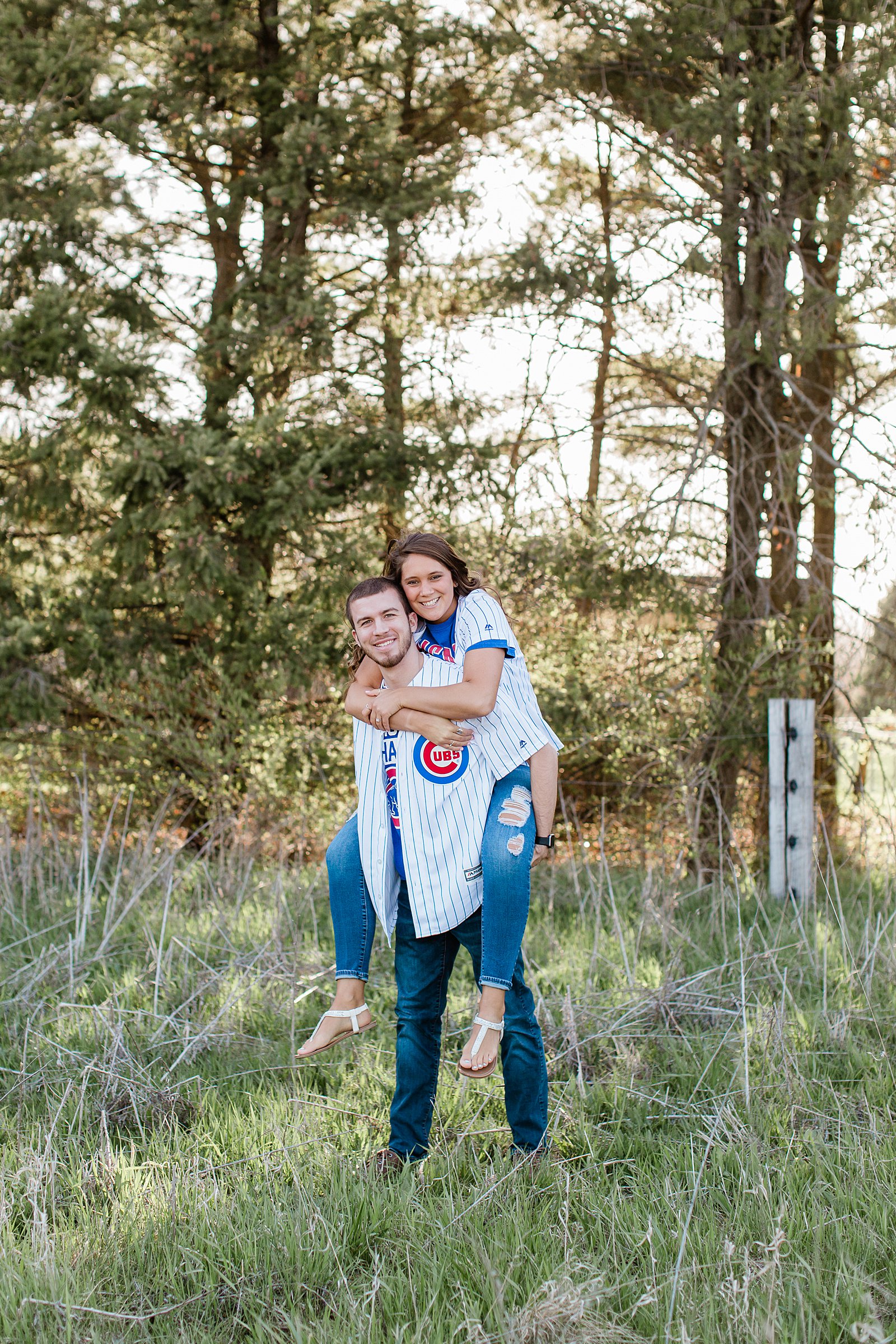 Guy giving his fiancé a piggy back ride and they are both smiling at the camera for their engagement photos. Photo is outside in the tall grass in Normal, Illinois. 